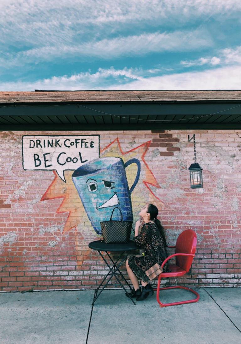 In this image, we see a woman sitting in front of a brick wall. Thoughtful, she looks at a graffiti on which is written "Drink Coffee Be Cool"